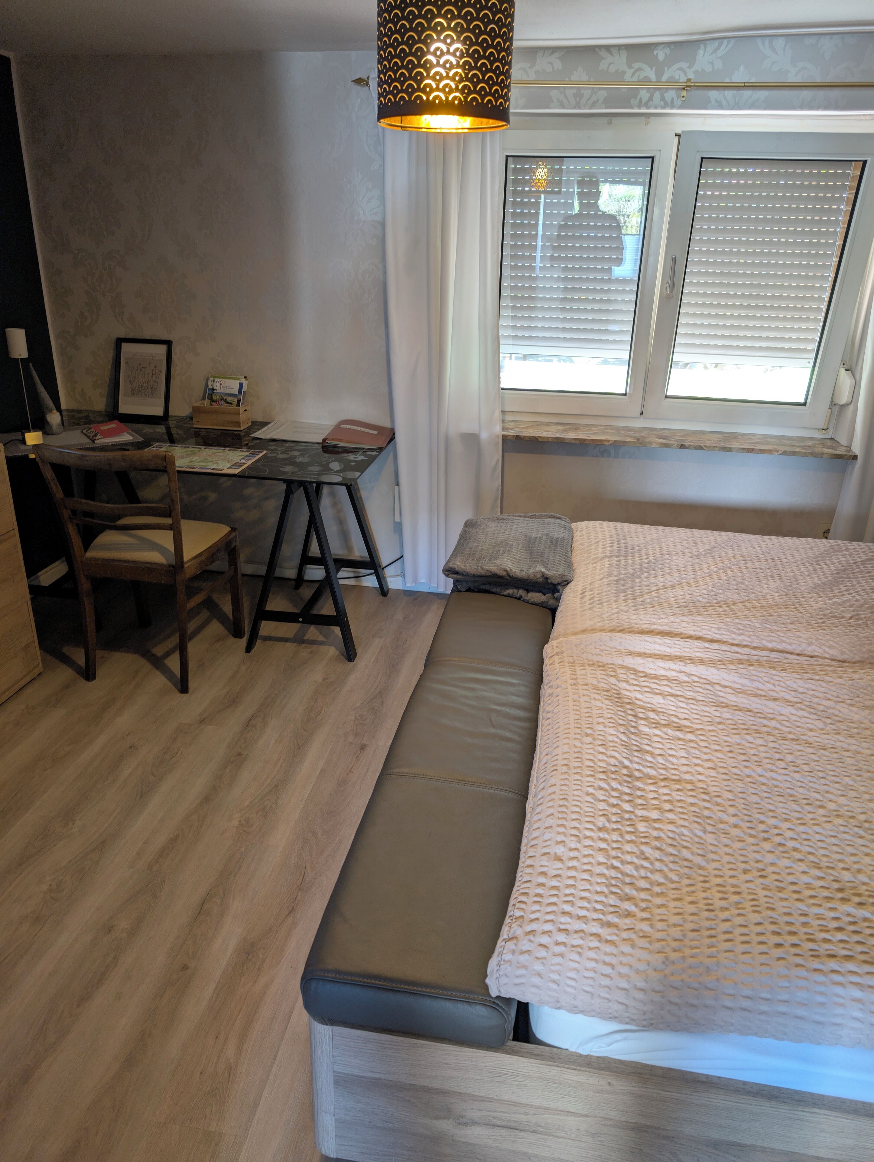 A compact room with a bed, a desk, and a chair. There is a window with blinds, a hanging light fixture, and a framed picture on the wall.