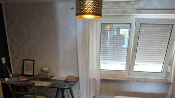 A compact room with a bed, a desk, and a chair. There is a window with blinds, a hanging light fixture, and a framed picture on the wall.