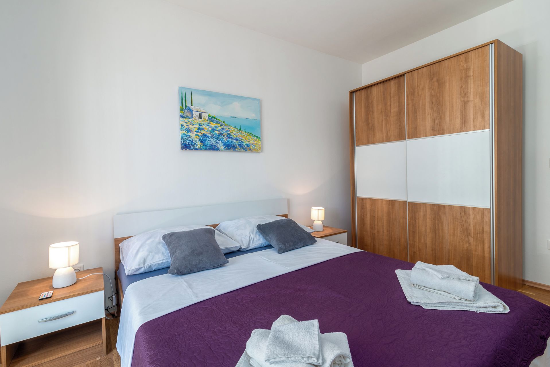 Et hotelværelse med seng, natbord, lampe, et billede og et klædeskab.