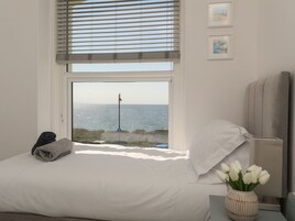 Un dormitorio con cama, ventana con vista al mar, lámpara y un florero con flores.
