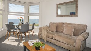 Uma sala de estar com um sofá bege, uma mesa de centro com revistas e vista para o mar através de grandes janelas.