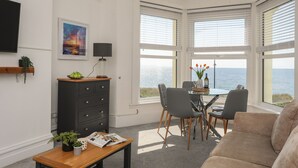 Uma sala de estar moderna com uma janela em arco com vista para o mar, uma área de jantar com mesa e cadeiras, e um armário preto com gavetas.