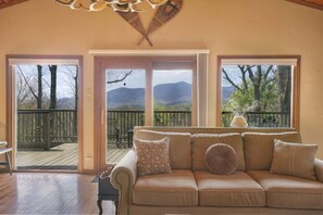 A cozy living room with a beige sofa, a wooden deck, and a view of mountains through large windows.