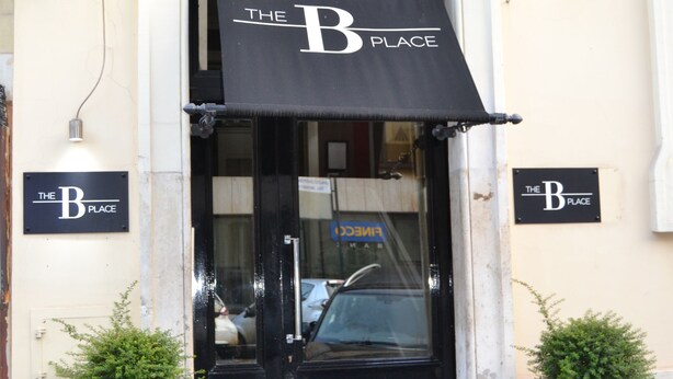 A building entrance with a black awning displaying "The B Place" sign, two potted plants, and a black door. - The B Place Ezio (Rome)