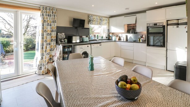 A modern kitchen with white cabinets, a dining table with a patterned tablecloth, and a fruit bowl. - Millbrook Cottage (Dalbeattie)