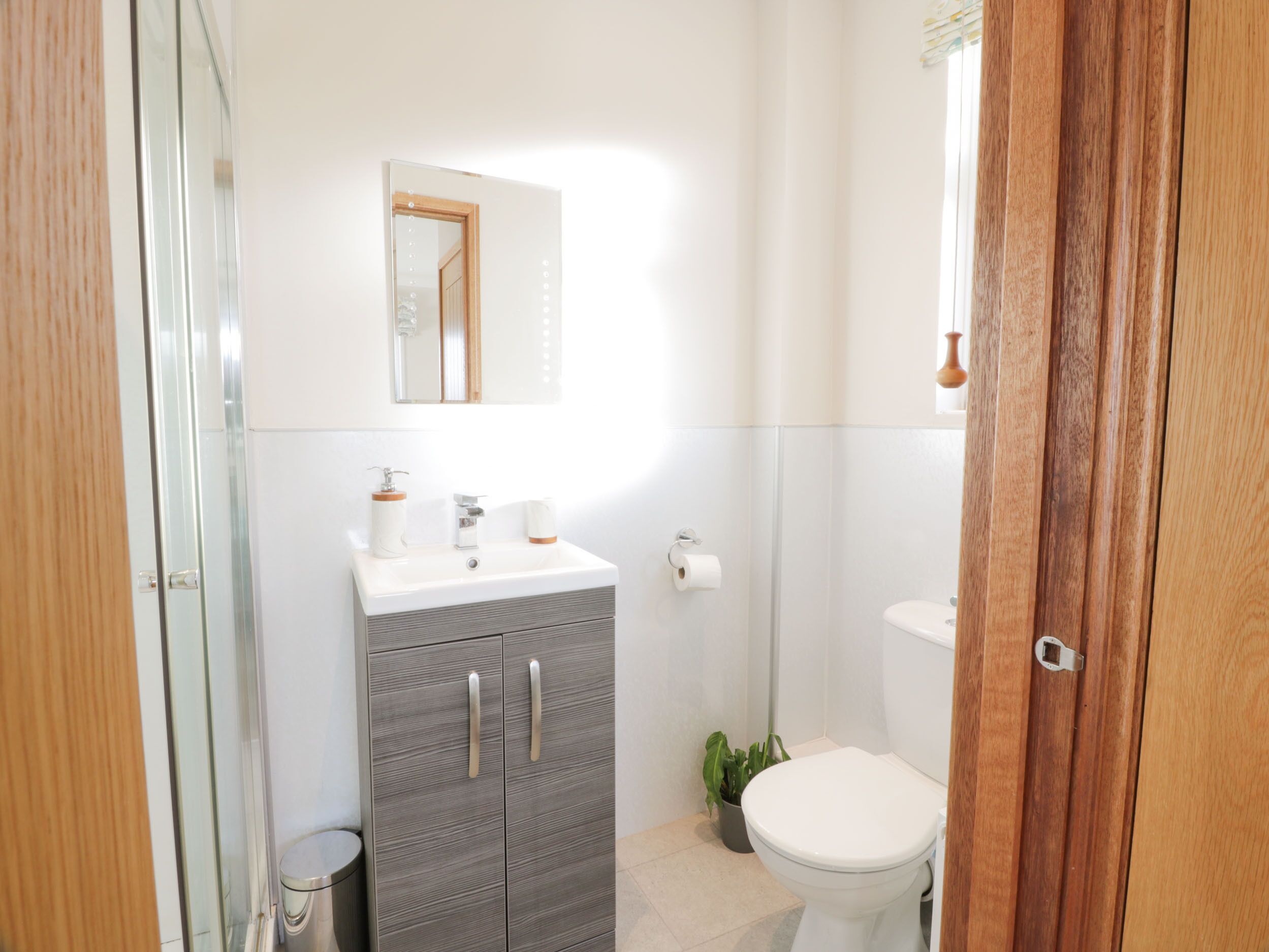 Une salle de bain moderne avec un lavabo blanc, un miroir et des toilettes. On y trouve une petite poubelle et une plante en pot.
