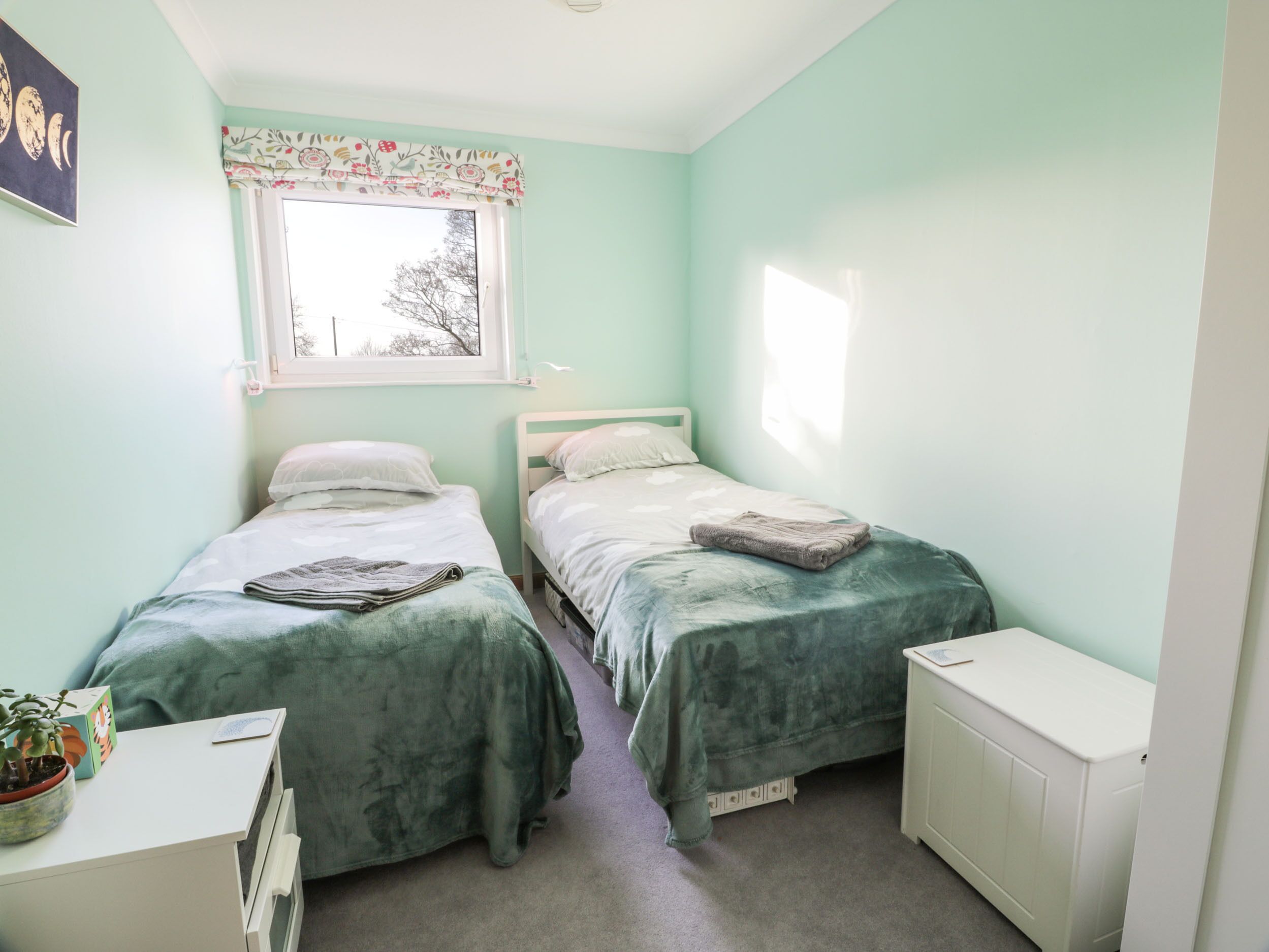 Un dormitorio pequeño y luminoso con dos camas individuales, un radiador blanco, un escritorio con una planta y una ventana con cortinas estampadas.
