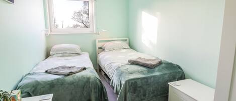 Un dormitorio pequeño y luminoso con dos camas individuales, un radiador blanco, un escritorio con una planta y una ventana con cortinas estampadas.