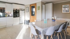 A modern kitchen with white cabinets, a built-in oven, and a dining area with a table and chairs. - Millbrook Cottage (Dalbeattie)