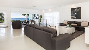 Una sala de estar moderna con un amplio sofá seccional gris, una mesa de centro blanca y un sofá marrón oscuro. Hay un ventanal con vista al océano y un balcón con barandilla.