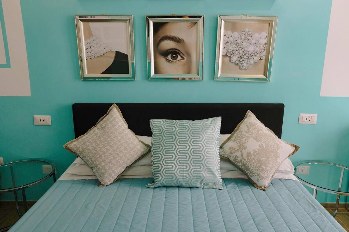A bed with a teal quilt, three patterned pillows, and three framed pictures on the wall.