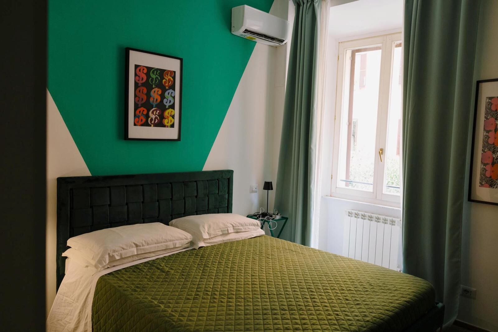 Una cama con colcha verde, cabecera negra y almohadas blancas en una habitación con pared verde y ventana con cortinas.