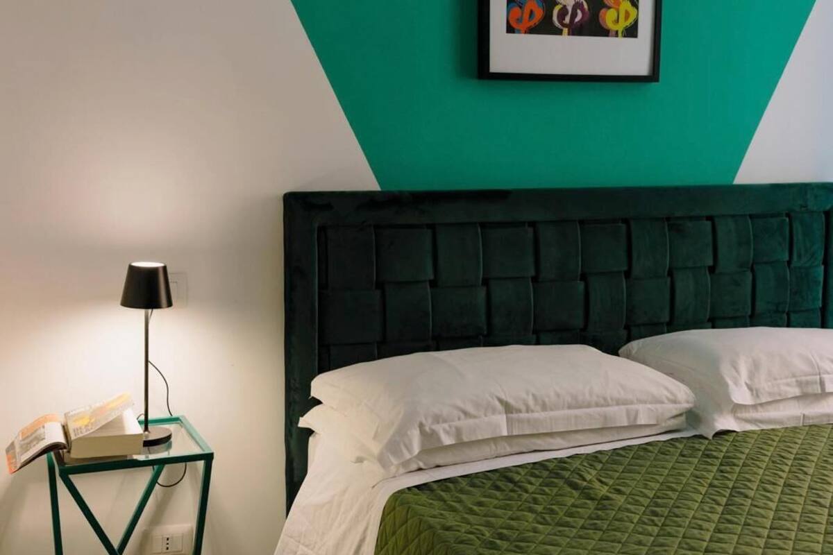 A bedroom with a green wall, a framed artwork of dollar signs, a black headboard, and a green quilted bedspread.