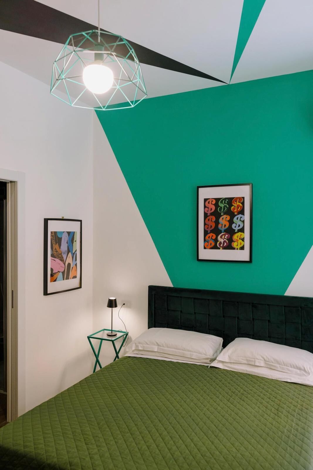 A bedroom with a green quilted bedspread, a geometric pendant light, and a framed artwork of dollar signs on the wall.