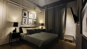 Un dormitorio con cama, dos mesitas de noche, una mesita auxiliar, televisión montada en la pared y un radiador.
