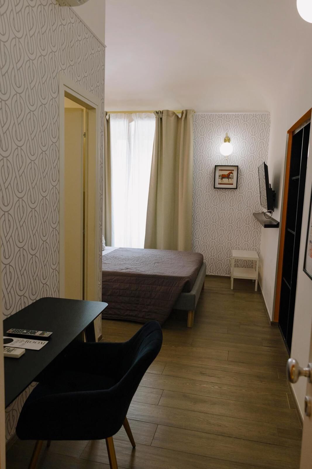 A small, compact room with a bed, a desk, and a chair. There is a window with curtains, a framed picture on the wall, and a small table with a television.