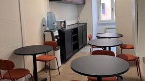 Una cocina moderna con armarios empotrados, microondas y lavabo. Varias mesas redondas y sillas naranjas dispuestas para comer.