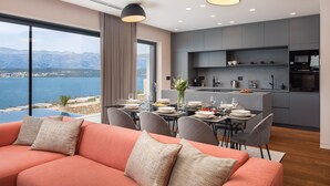A modern living room with a sofa and dining area, a kitchen with grey cabinets, and a view of the sea.