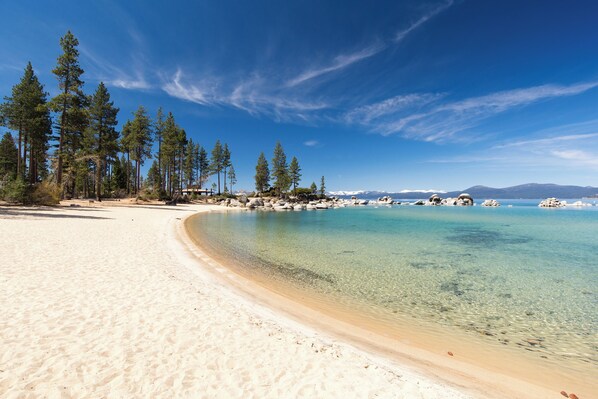 A sandy beach with clear turquoise water, surrounded by tall pine trees and distant mountains under a clear blue sky.