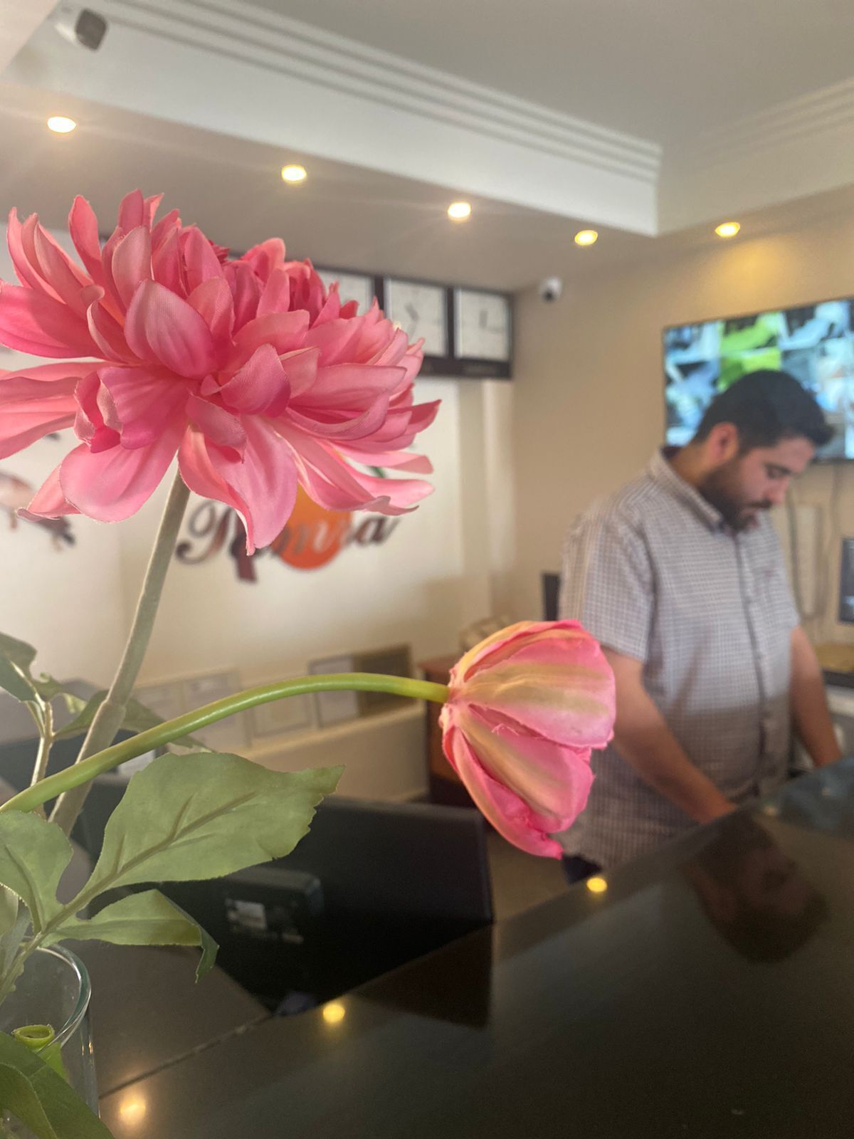 Eine rosa Blume in einer Vase auf einem Tisch, im Hintergrund arbeitet ein Mann an einer Theke.