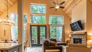 Interior acogedor de una cabaña con chimenea de piedra, ventilador de techo y grandes ventanales con vistas al bosque.