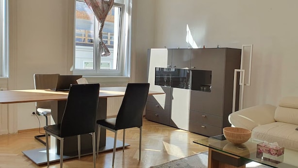 A modern living room with a wooden floor, a glass coffee table, a sofa, and a dining area with a table and chairs. - Servicierte Wohnung in der City (Vienna)