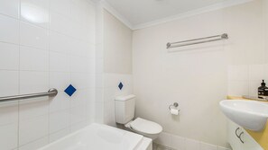 Une salle de bain avec des carreaux blancs, une baignoire, des toilettes, un distributeur de savon liquide et un porte-serviettes.