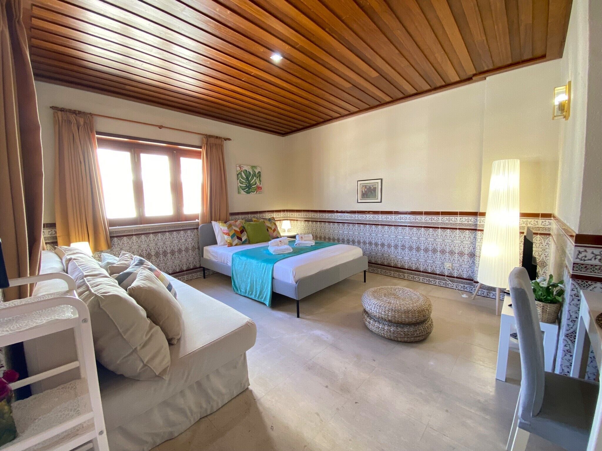 A hotel room with a wooden ceiling, a bed with a blue blanket, a sofa, a small round ottoman, and a chair.
