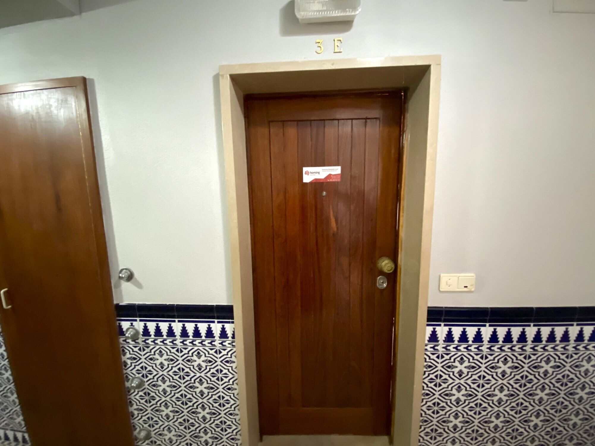 A wooden door with a sign above it, a tiled wall with a geometric pattern, and a light switch on the wall.