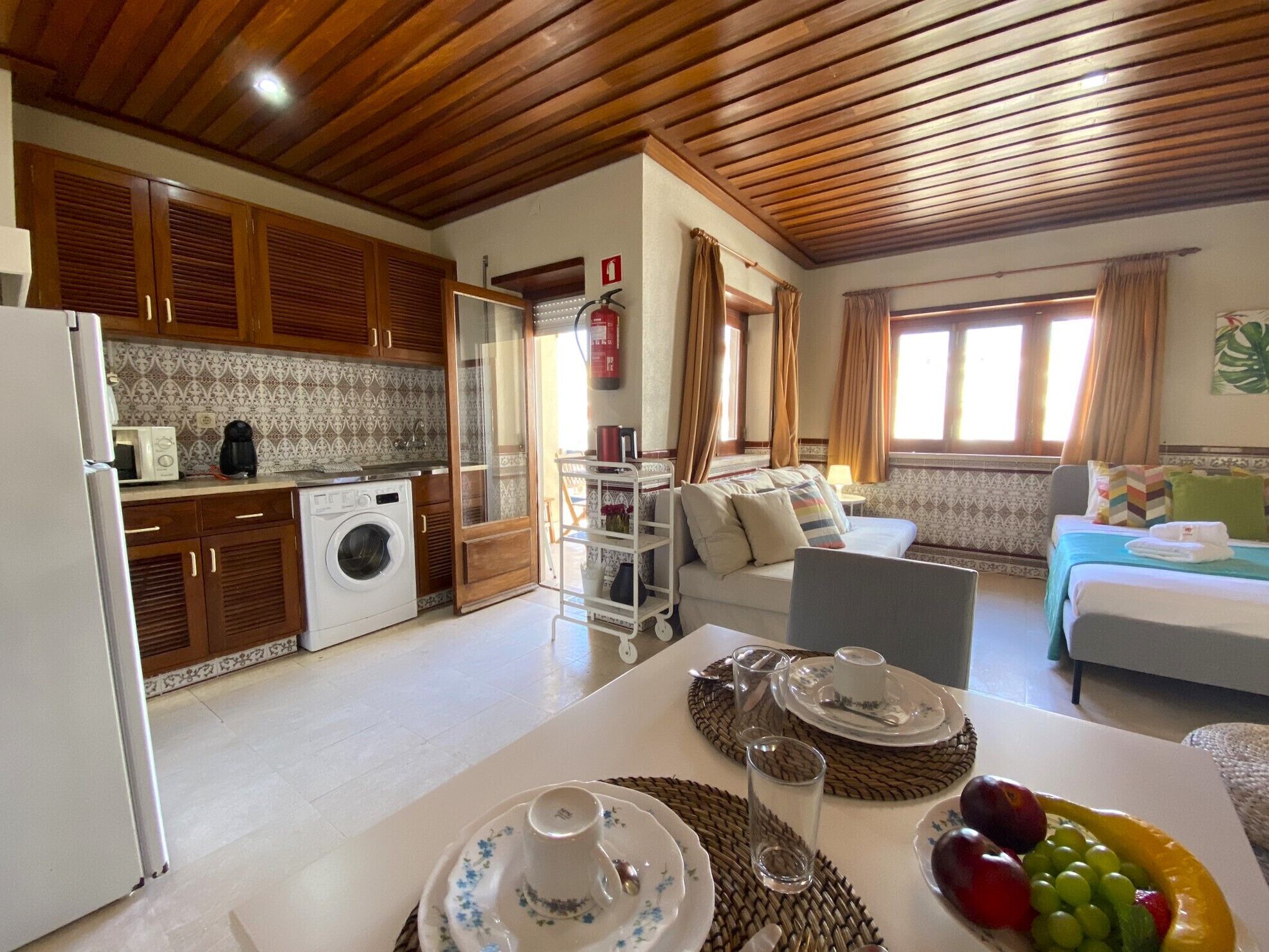 Quarto de hotel compacto com cozinha compacta, sofá, mesa de jantar com pratos e copos, e uma cesta de frutas.