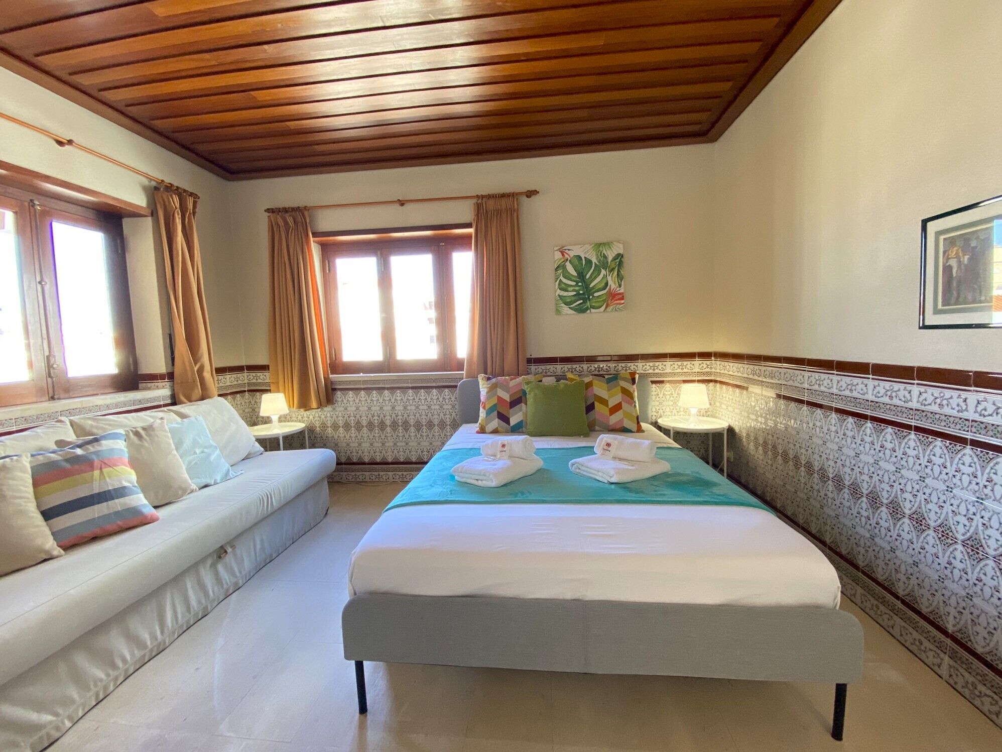 A hotel room with a bed, a sofa, a window with curtains, a wall with a plant, and a patterned wall border.