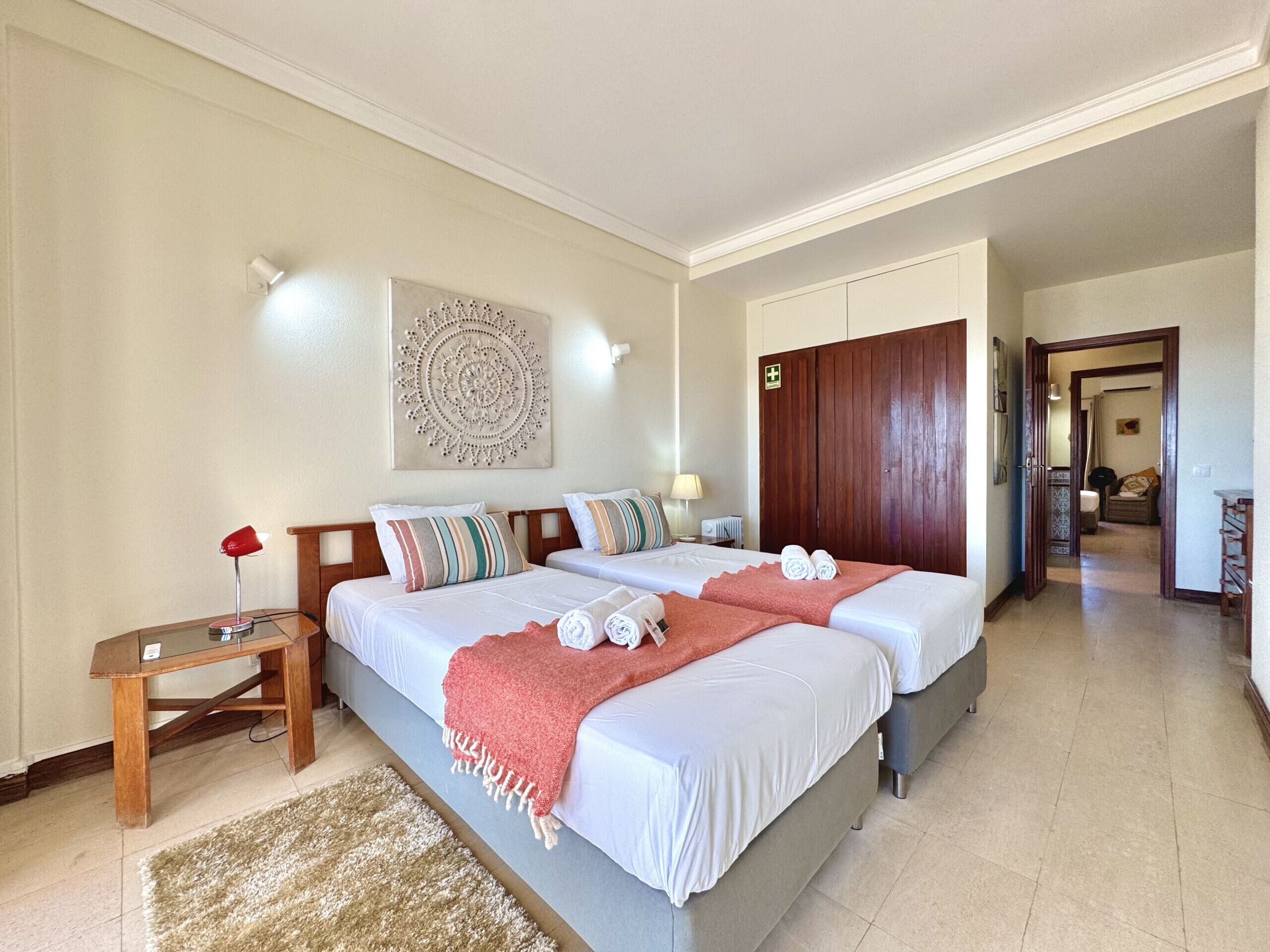 A hotel room with two beds, a wooden nightstand, a red lamp, a wall-mounted artwork, and a door leading to another room.