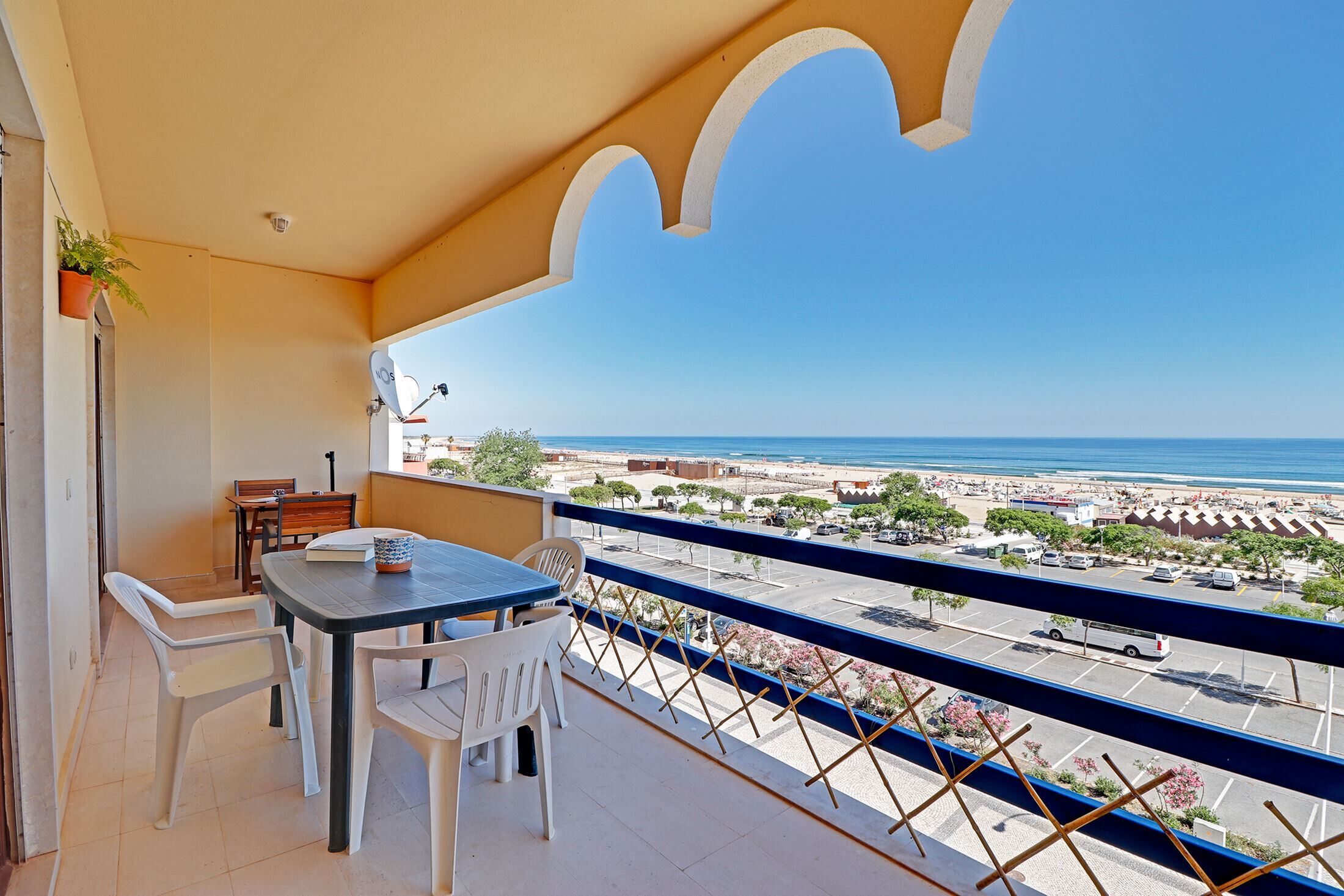 A balcony with a table and chairs, overlooking a beach and parking area.