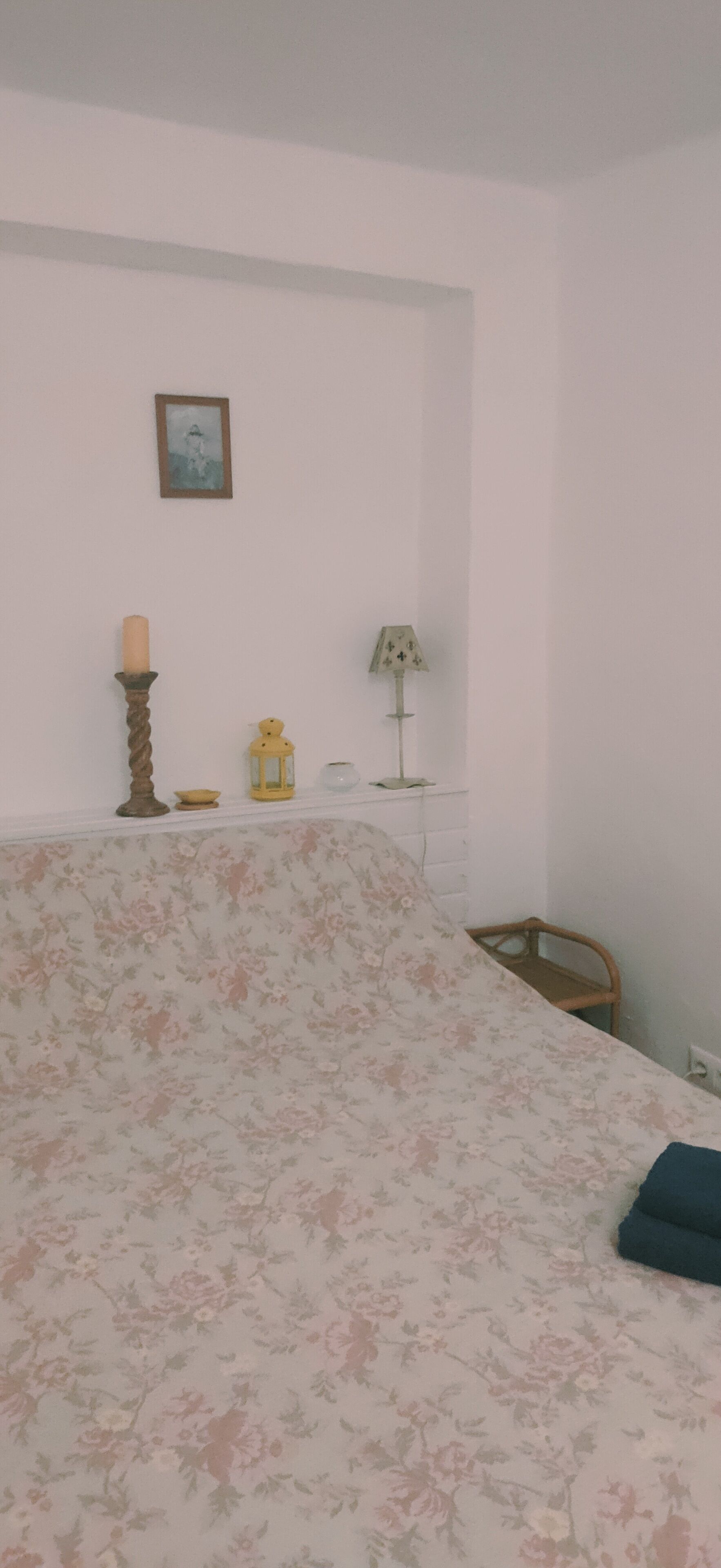 A bedroom with a floral bedspread, a bedside table with a lamp, a candle, and a small decorative item.