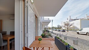 A balcony with a wooden table and chairs, a potted plant, and a view of a street with cars and buildings. - Tavira Formosa BAY 1 by Homing (Tavira)