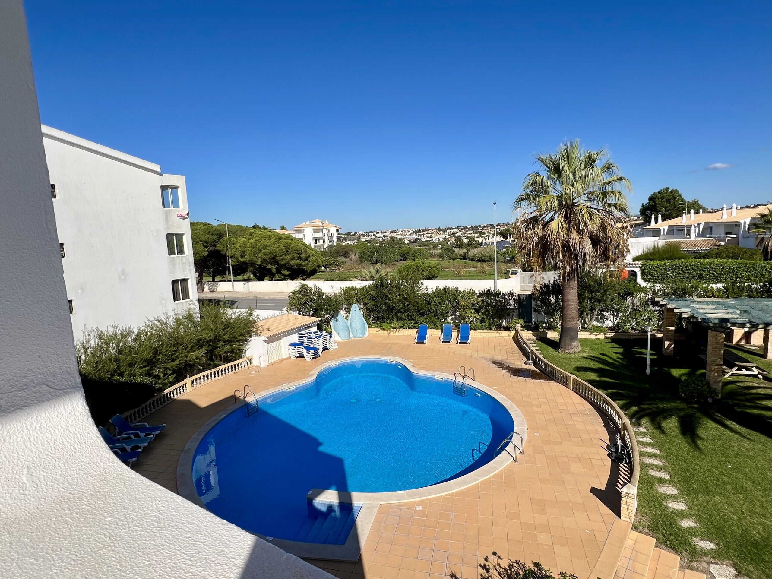 En swimmingpool med vandrutsjebaner, omgivet af fliseterrasse, palmer og udsigt til boligområde.