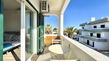 En balkon med seng, bord og stole. Klimaanlæg og udsigt til omkringliggende bygninger.
