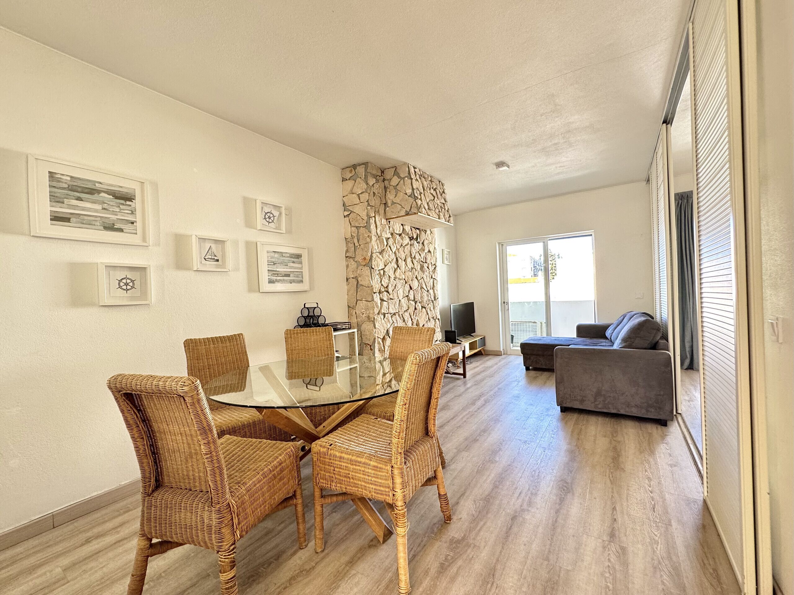 A modern living room with a glass-top dining table surrounded by wicker chairs, a stone accent wall, and a grey sofa.