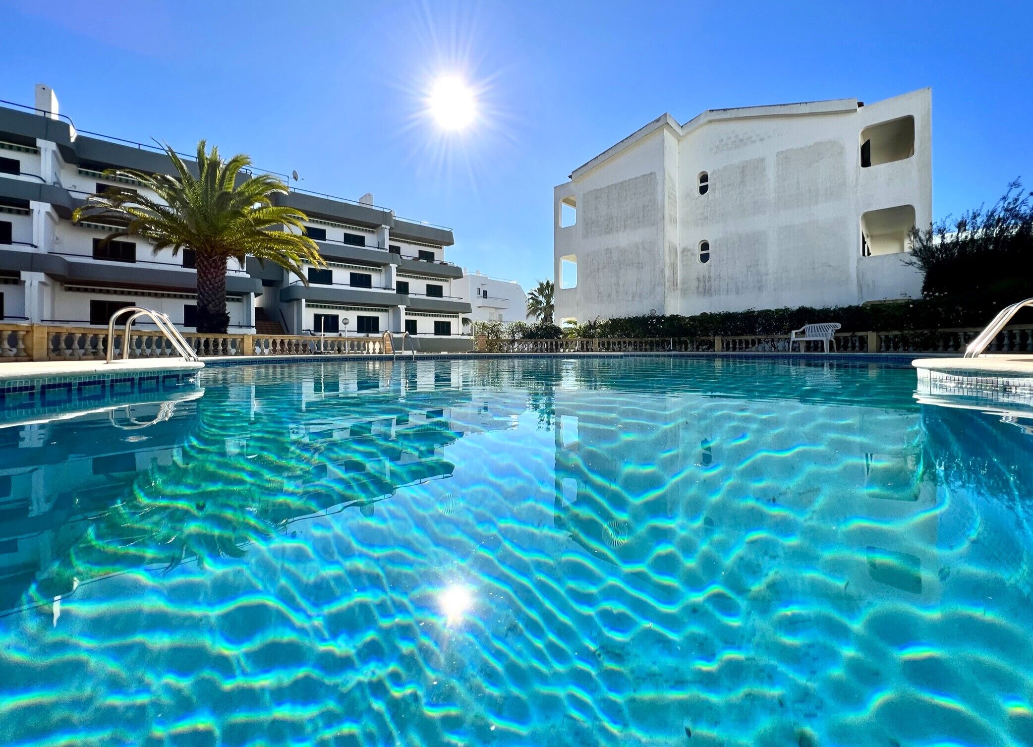 A clear swimming pool with a sun reflecting on the water, surrounded by a modern building with balconies and a palm tree.