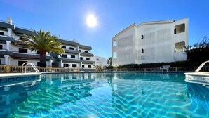 A clear swimming pool with a sun reflecting on the water, surrounded by a modern building with balconies and a palm tree.
