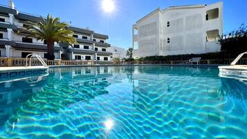 A clear swimming pool with a sun reflecting on the water, surrounded by a modern building with balconies and a palm tree.