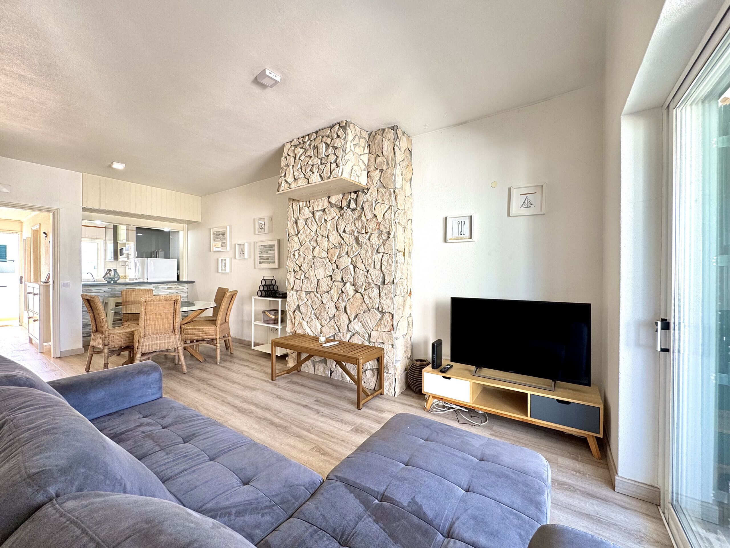 A modern living room with a stone accent wall, a flat-screen TV, and a dining area with wicker furniture.