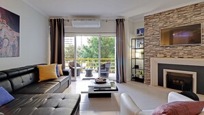 A modern living room with a black leather sofa, a white armchair, and a coffee table with a bowl of fruit. There is a fireplace and a TV mounted on a stone wall. - Vilamoura Design With Pool by Homing (Loulé)