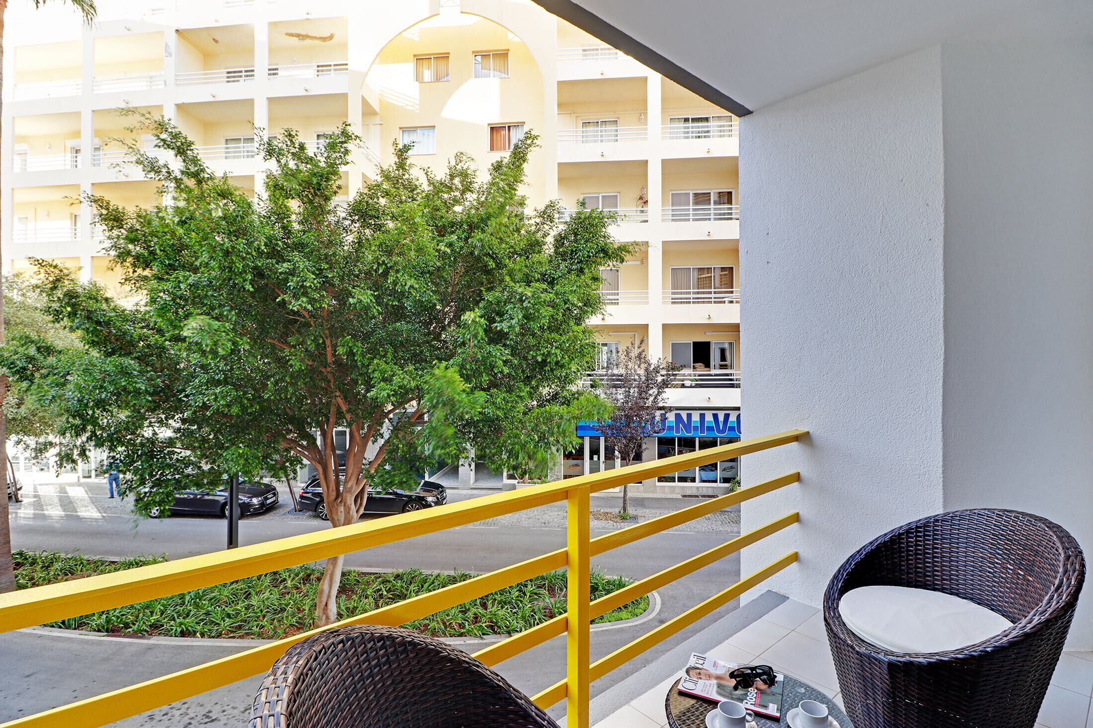 A balcony with wicker chairs, a yellow railing, and a view of a building with multiple balconies and parked cars.