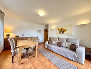 A living room with a wooden dining table, wicker chairs, a grey sofa with patterned cushions, a rug, and a painting on the wall. - Pera Sea View With Pool by Homing (Silves)