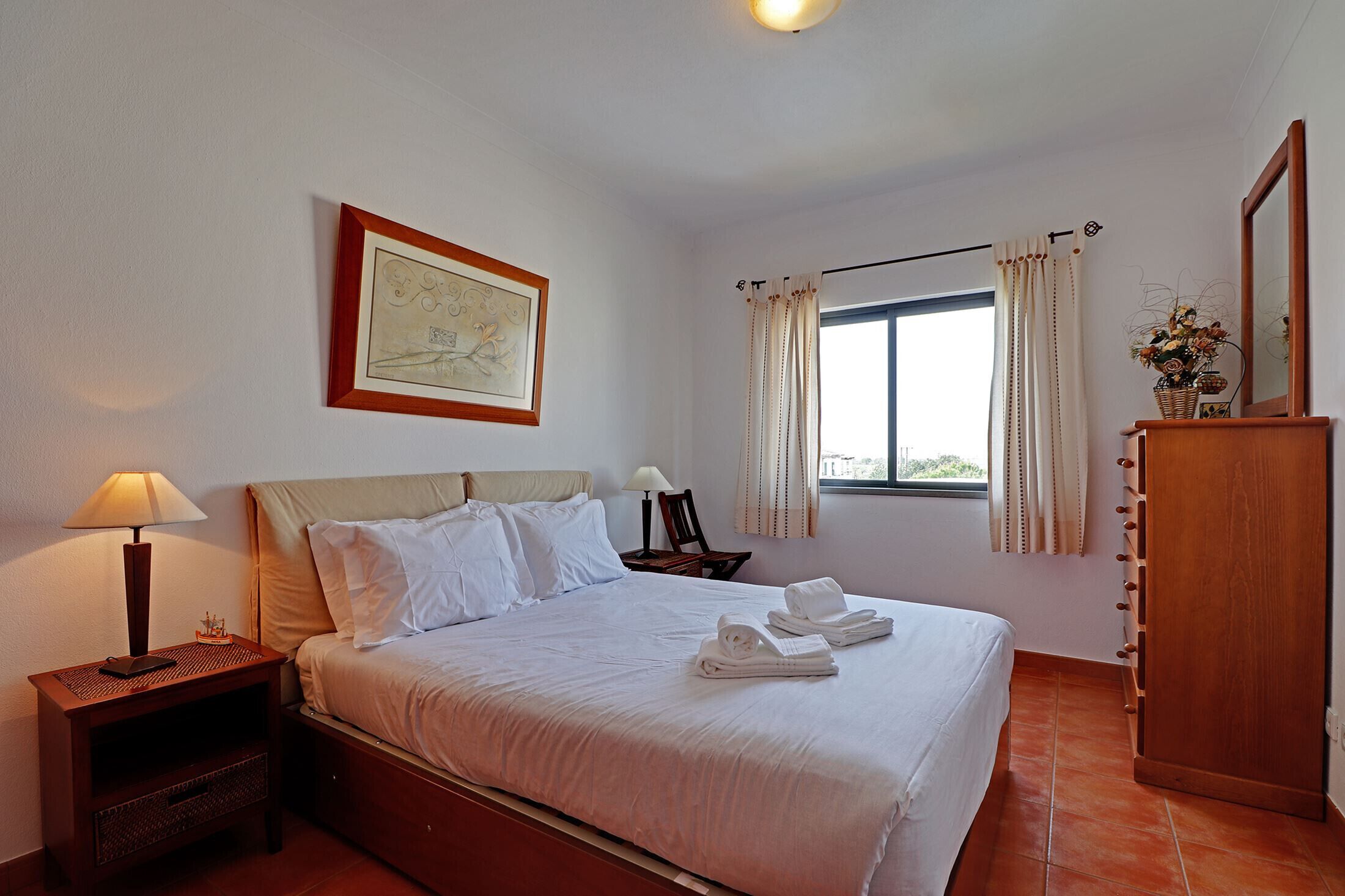 Una cama bien hecha con sábanas blancas, una lámpara de noche, un cuadro en la pared, una ventana con cortinas y un armario de madera.