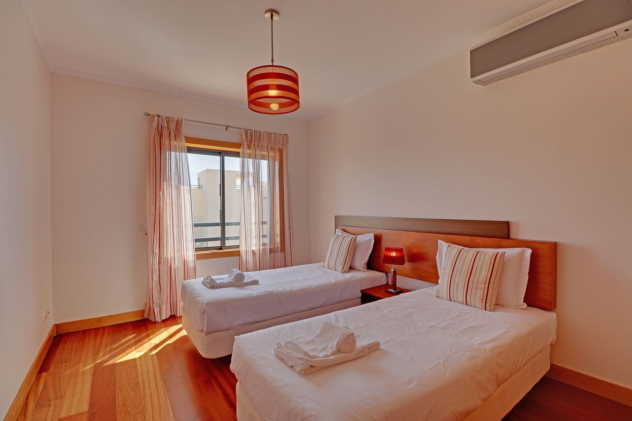 A hotel room with two beds, a window with curtains, a wooden floor, and a red lamp on the nightstand.