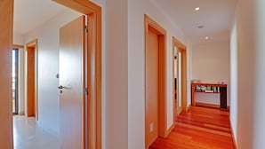 Um corredor com portas e soalho de madeira, que dá para um quarto com uma estante de livros.