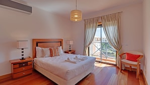 Quarto de hotel com uma cama grande, duas mesas de cabeceira, uma cadeira, uma janela com cortinas e vista para prédios.