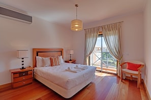 Ein Hotelzimmer mit einem großen Bett, zwei Nachttischen, einem Sessel, einem Fenster mit Vorhängen und Blick auf Gebäude.
